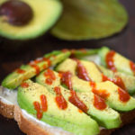Sliced avocado on bread with drizzle of hot sauce with cut avocado in the background