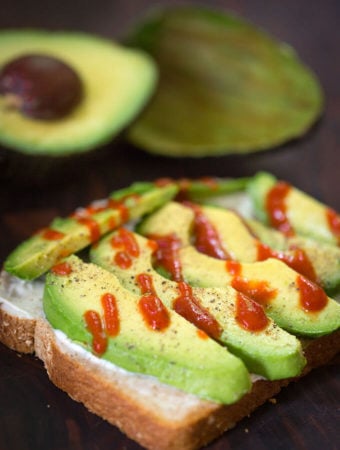 Sliced avocado on bread with drizzle of hot sauce with cut avocado in the background