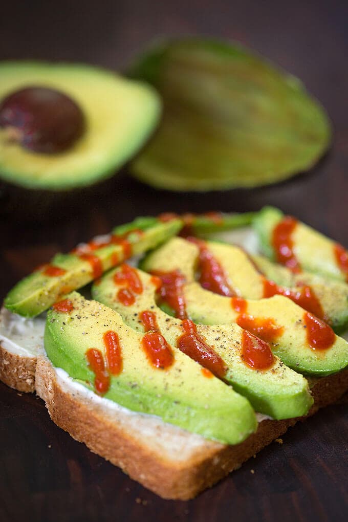 Sliced avocado on bread with drizzle of hot sauce with cut avocado in the background