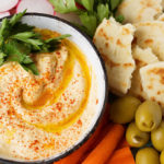 Hummus in a small white bowl on platter filled with carrot sticks, sliced radishes, torn flat bread and green onions