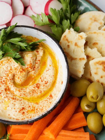 Hummus in a small white bowl on platter filled with carrot sticks, sliced radishes, torn flat bread and green onions