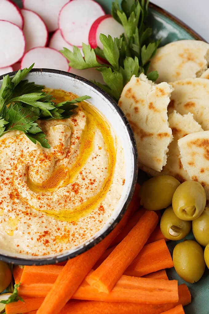 Hummus in a small white bowl on platter filled with carrot sticks, sliced radishes, torn flat bread and green onions