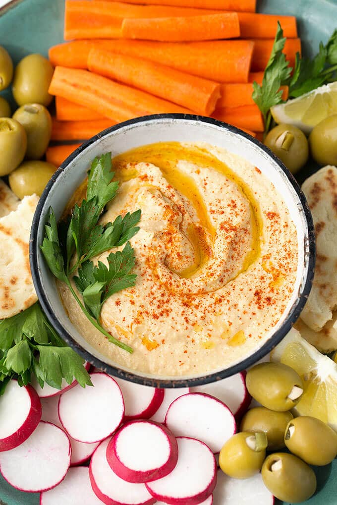  Hummus in a small white bowl on platter filled with carrot sticks, sliced radishes, torn flat bread and green onions