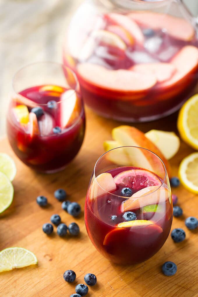 Two Fruity Red Sangria in glasses on a wooden board with a pitcher, blueberries and lemon slices in the background