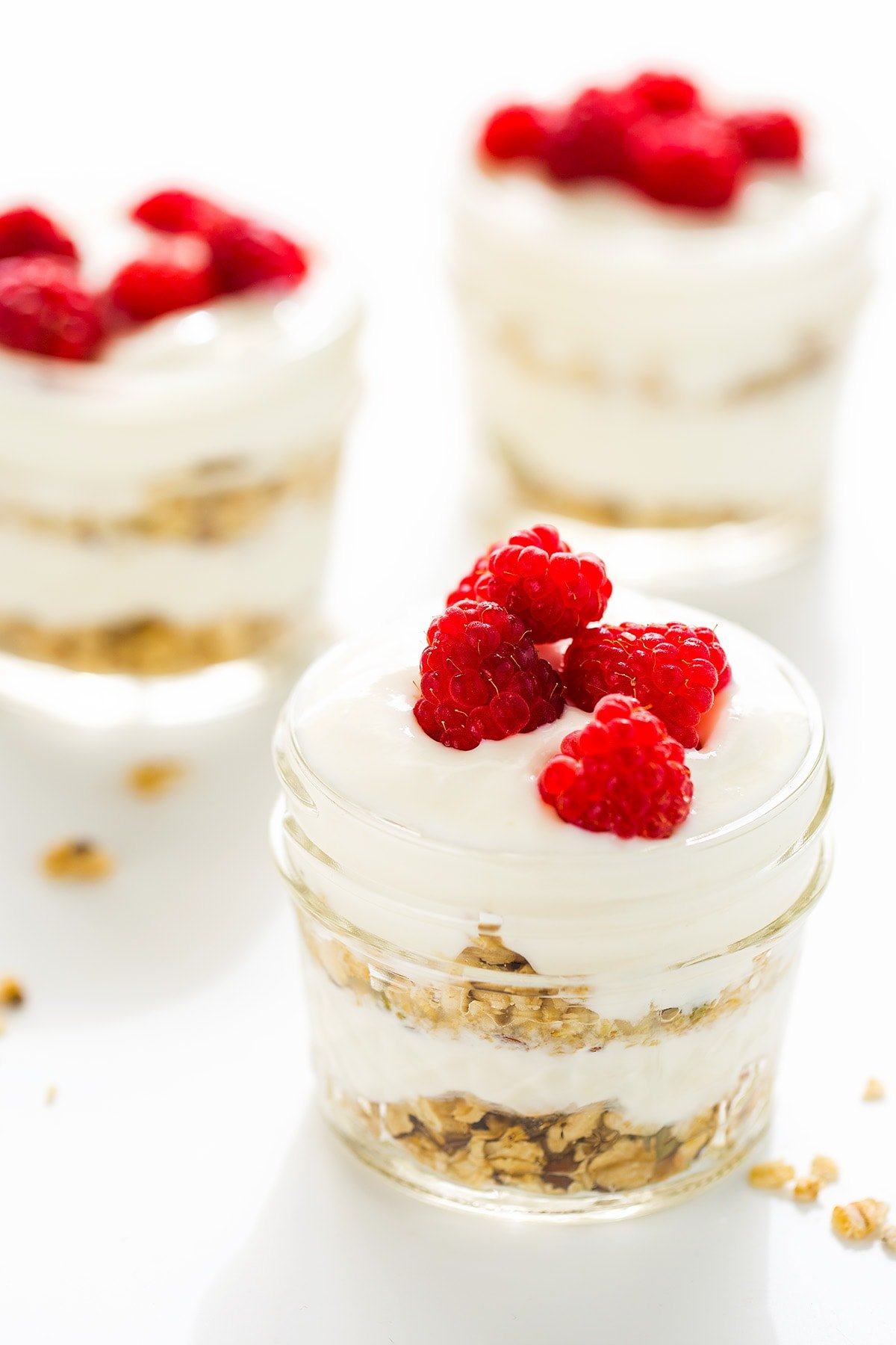 three glass jars layered with granola and yogurt topped with raspberries