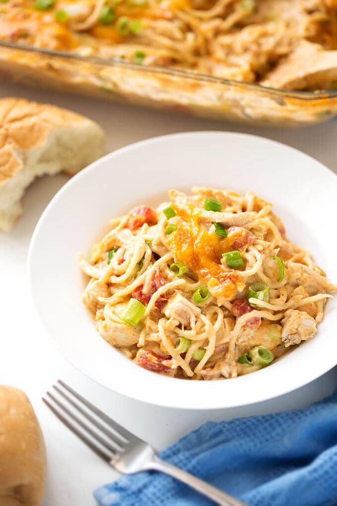 Creamy Baked Chicken Spaghetti in white bowl next to fork and blue napkin with casserole dish in the background