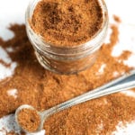 Easy Taco Seasoning in small glass jar with some spilled on white background with small silver measuring spoon