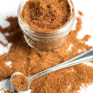 Easy Taco Seasoning in small glass jar with some spilled on white background with small silver measuring spoon