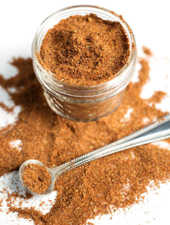 Easy Taco Seasoning in small glass jar with some spilled on white background with small silver measuring spoon