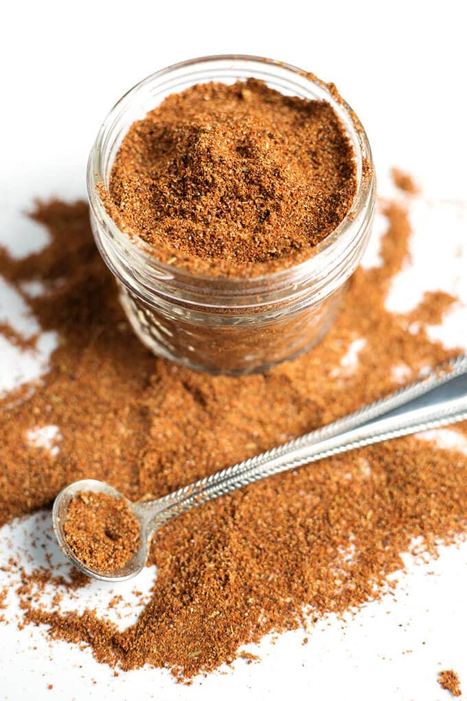 Easy Taco Seasoning in a small glass jar some spilled on white surface with small silver measuring spoon