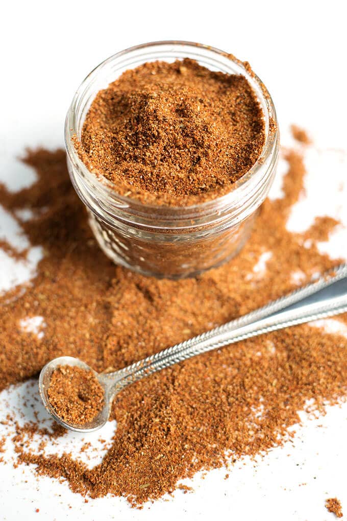 Easy Taco Seasoning in small glass jar with some spilled on white background with small silver measuring spoon