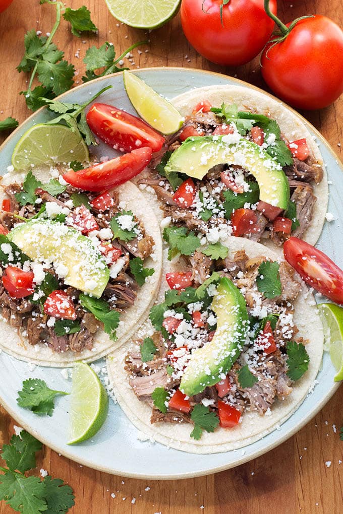 Three Instant Pot Pork Carnitas on corn tortillas on a white plate garnished with lime and tomato wedges