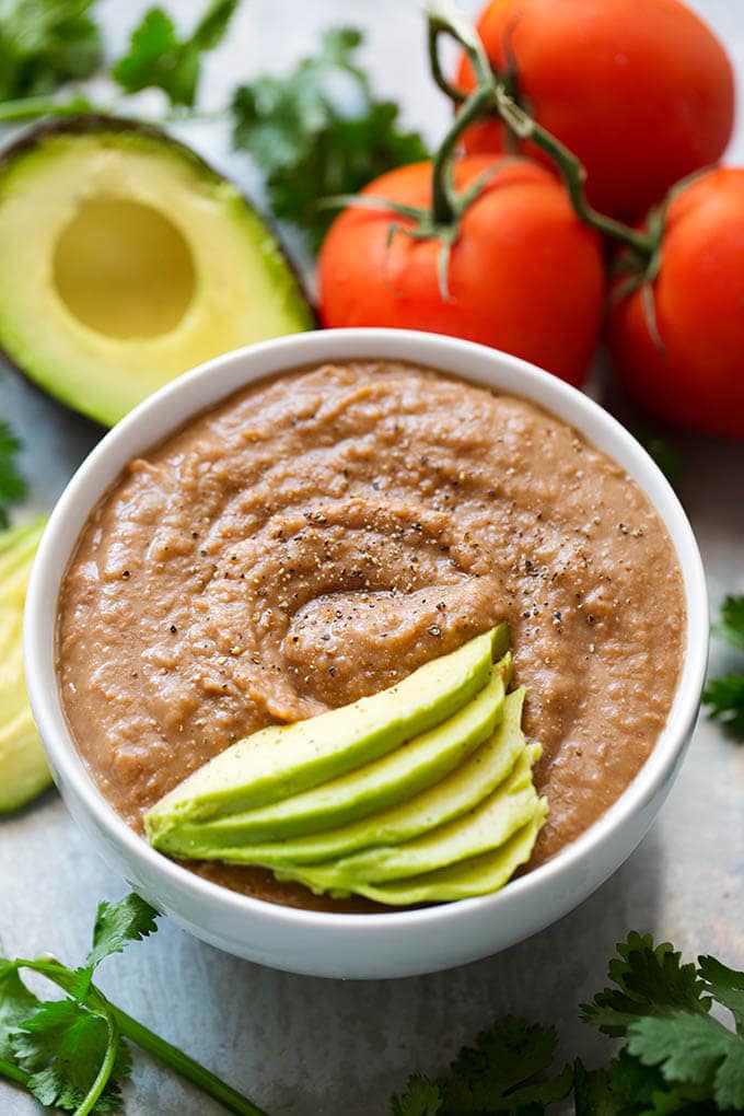 Instant Pot Refried Beans in small white bowl topped with thinly sliced avocado with avocado and tomatoes in the background