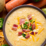 Loaded Broccoli Cheese & Potato Soup in a dark colored bowl with carrots and broccoli in the background