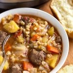 Instant Pot Beef Barley Vegetable Soup in a white bowl with spoon next to two rolls