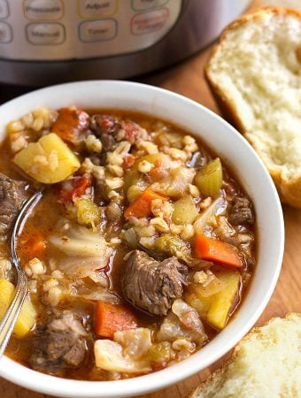 Instant Pot Beef Barley Vegetable Soup in a white bowl with spoon next to two rolls