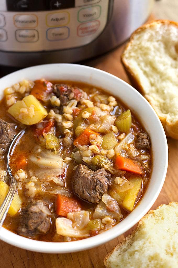 Instant Pot Beef Barley Vegetable Soup in a white bowl with spoon next to two rolls