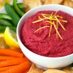Red Beet Hummus in a white bowl surrounded by pita chips carrot sticks green pepper sticks and lemon wedges