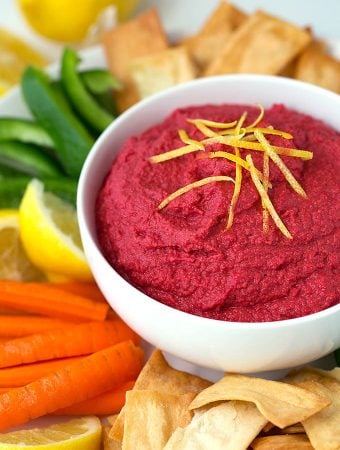 Red Beet Hummus in a white bowl surrounded by pita chips carrot sticks green pepper sticks and lemon wedges