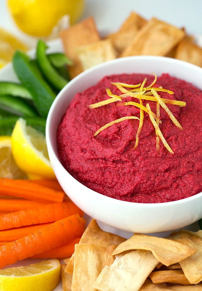 Red Beet Hummus in a white bowl surrounded by pita chips carrot sticks green pepper sticks and lemon wedges