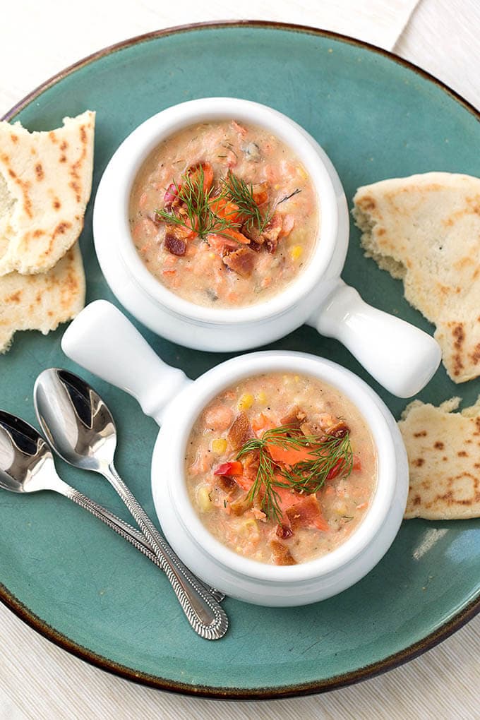 Two white bowls of Salmon Chowder on turquoise plate with two silver spoons and torn flat bread