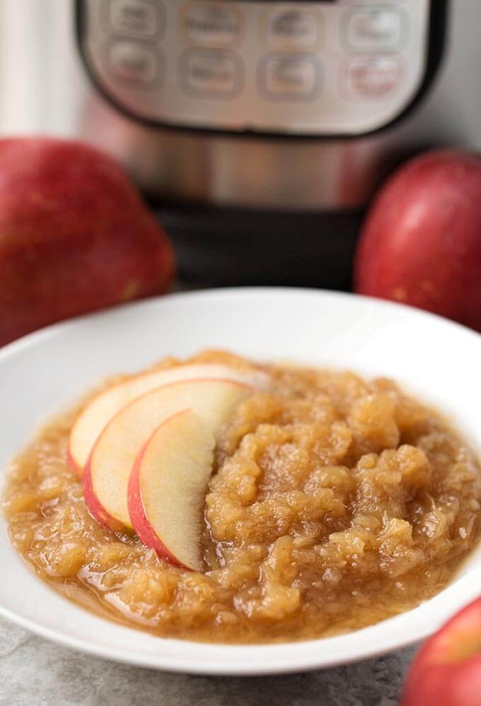 Applesauce in a shallow white bowl with sliced apples with pressure cooker and apples in background