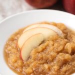 Applesauce in a shallow white bowl with sliced apples with pressure cooker and apples in background