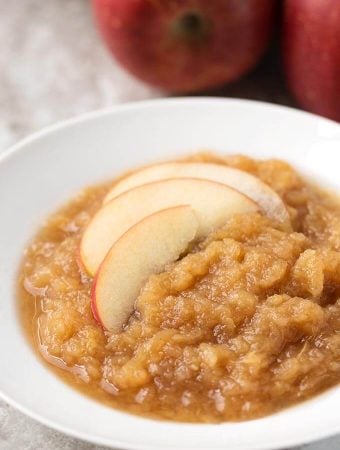 Applesauce in a shallow white bowl with sliced apples with pressure cooker and apples in background