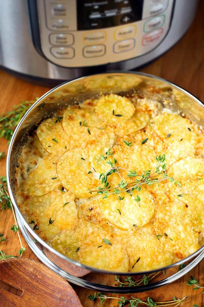 Cheesy Scalloped Potatoes in a round pan on a wooden board with pressure cooker in background