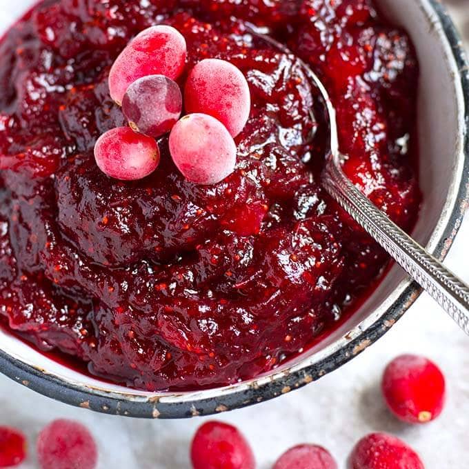 Cranberry Sauce in a white bowl with black rim surrounded by frosted cranberries and a spoon