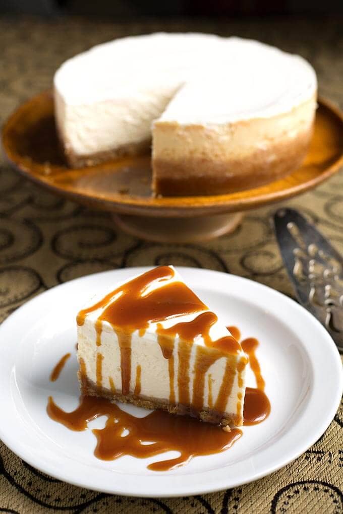 Slice of New York Cheesecake drizzled with caramel sauce on white plate with rest of cake in the background