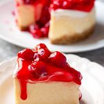 Slice of New York Cheesecake covered with cherry topping on a white plate with the rest of the cake in the background