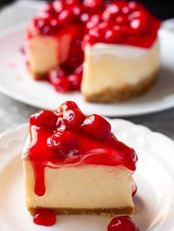 Slice of New York Cheesecake covered with cherry topping on a white plate with the rest of the cake in the background