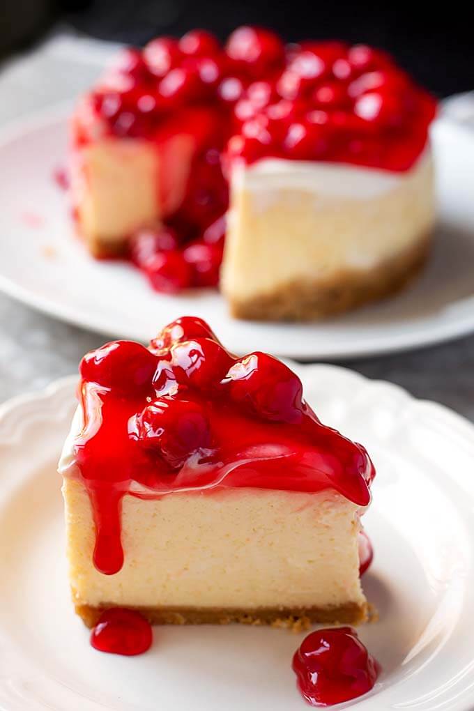 Slice of New York Cheesecake covered with cherry topping on a white plate with the rest of the cake in the background