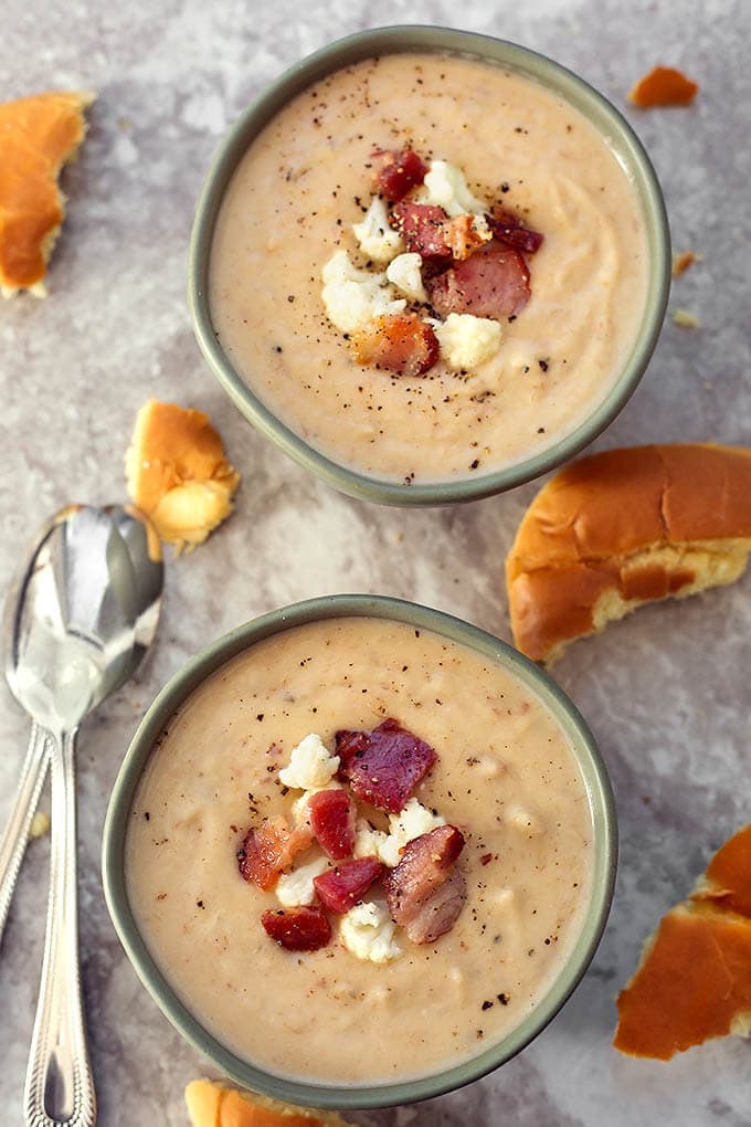 Two grey bowls of Cauliflower Bacon Chowder surrounded by torn bread and two silver spoons