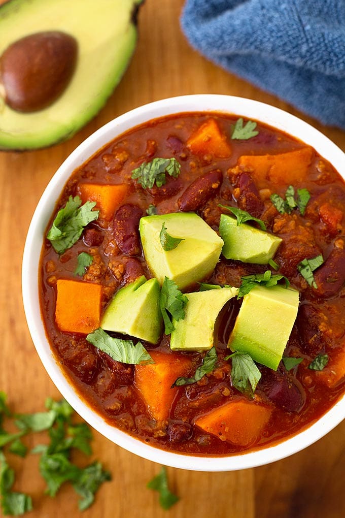 Sweet potato chili in a white bowl on wooden board garnished with chopped avocados