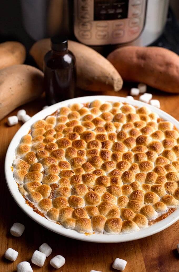 Candied Yams in a white bowl in front of raw sweet potatoes in front of a pressure cookers