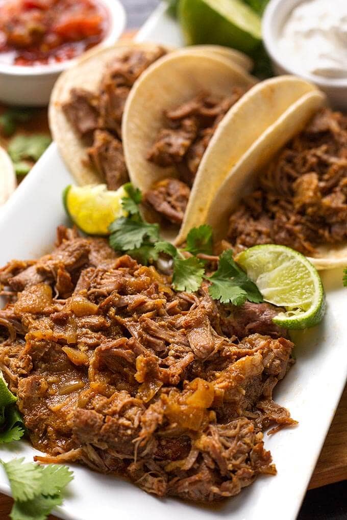 Close up of Pork Carnitas in small tortillas on a white platter garnished with lime and cilantro