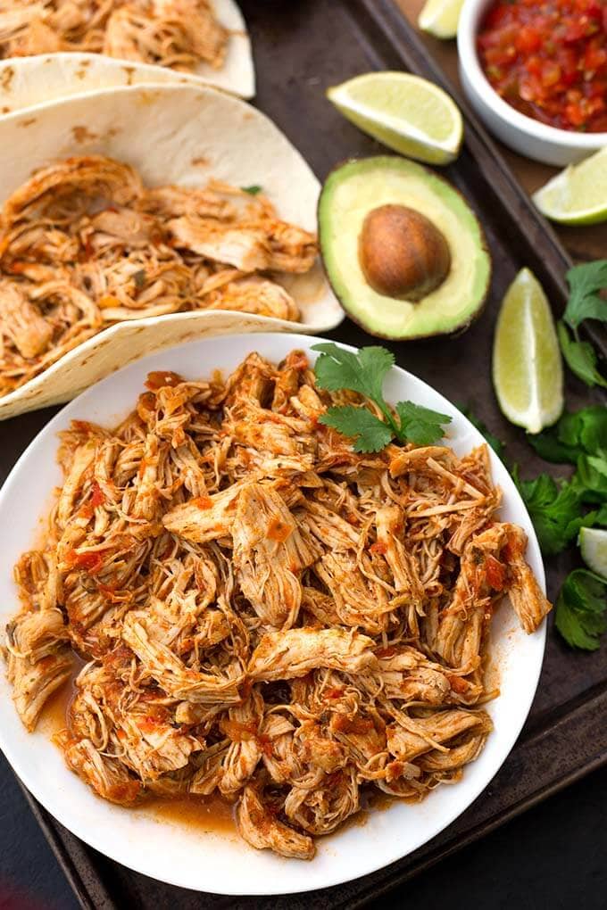 Salsa Chicken on a white plate next to salsa chicken in flour tortilla with halved avocado and limes in background