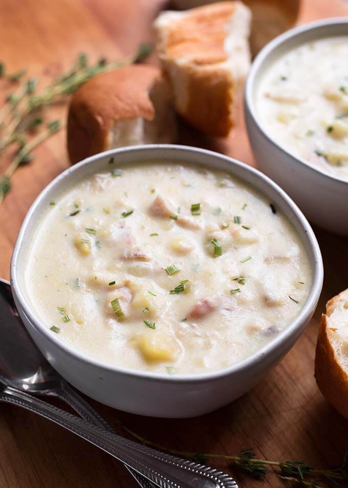 Two white bowls of Clam Chowder on a wooden board with bread rolls and silver spoons