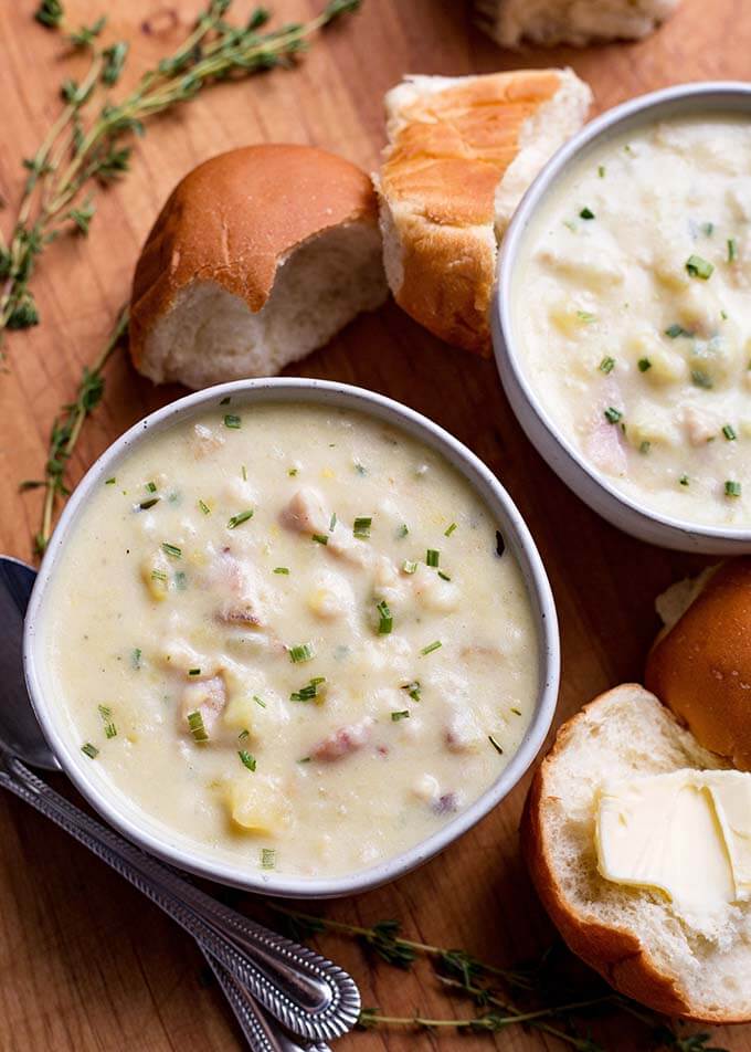 Two white bowls of Clam Chowder on a wooden board with bread rolls