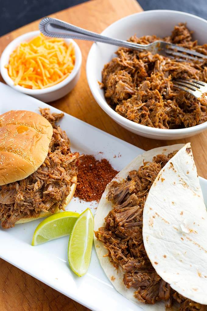 Pulled Pork on a hamburger bun next to pulled pork in flour tortilla on a rectangular white platter with bowl of pulled pork and knife in background