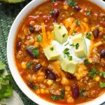 Taco Soup garnished with cubed avocado in a white bowl next to silver bowl on green mat