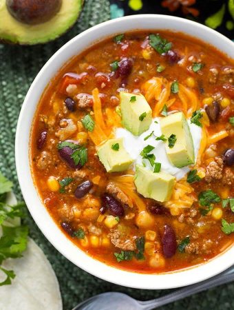 Taco Soup garnished with cubed avocado in a white bowl next to silver bowl on green mat