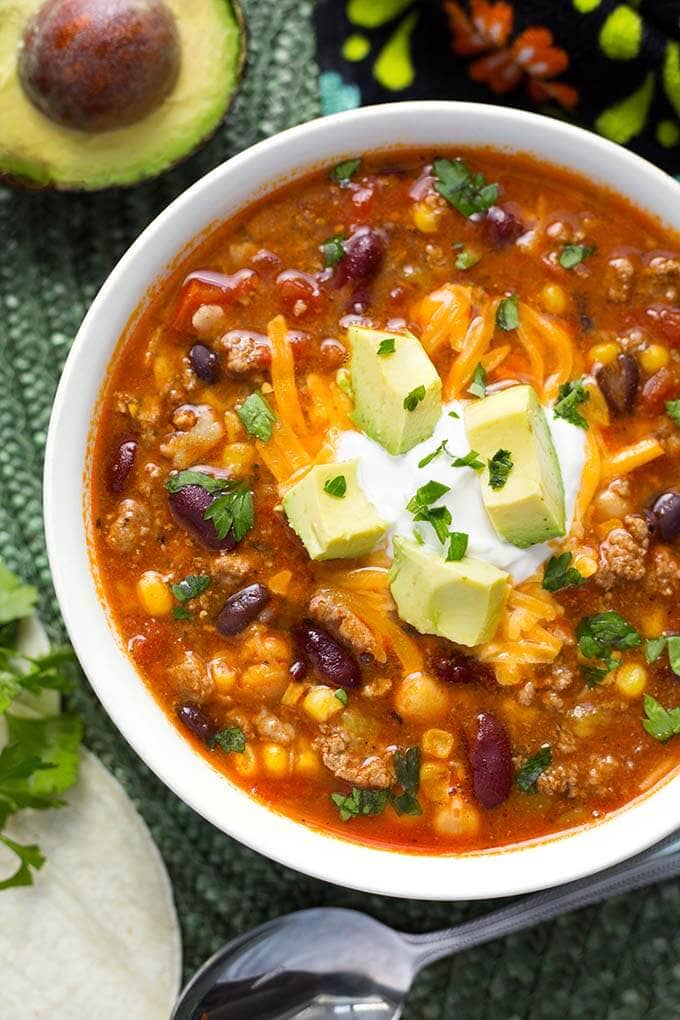 Taco Soup garnished with cubed avocado in a white bowl next to silver bowl on green mat 