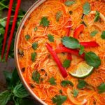 Red Curry Coconut Noodle Soup in a beige bowl next to red chopsticks and herbs