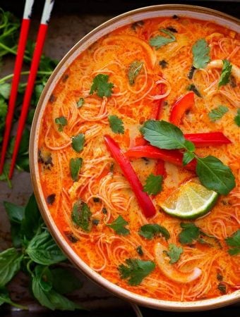 Red Curry Coconut Noodle Soup in a beige bowl next to red chopsticks and herbs