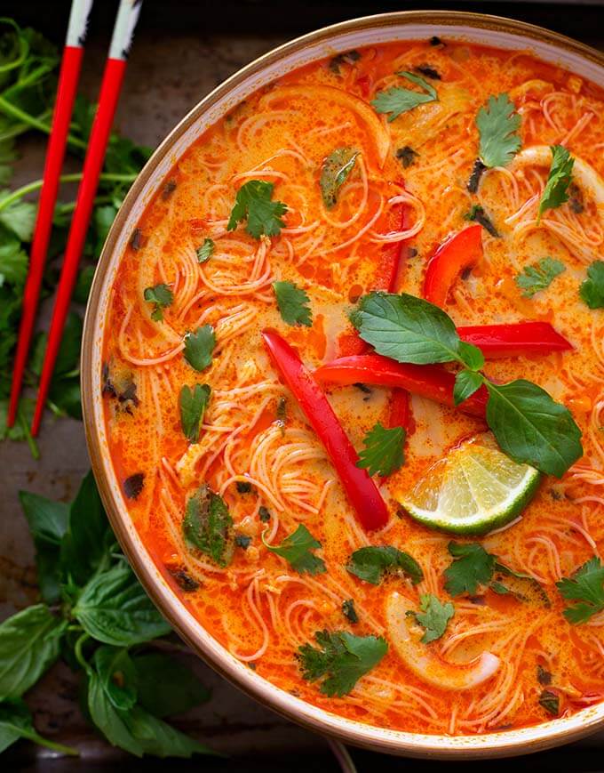 Red Curry Coconut Noodle Soup in a beige bowl next to red chopsticks and herbs