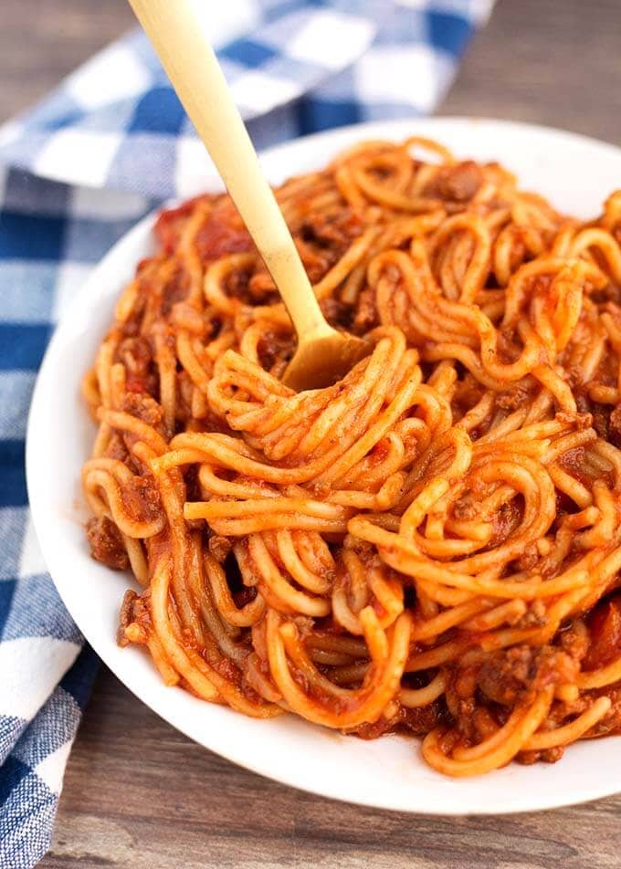 instant pot Spaghetti on a white plate with golden fork twisting noodles and gingham napkin on wooden board.