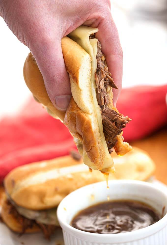 Hand dipping French Dip Sandwich in small white bowl of brown sauce